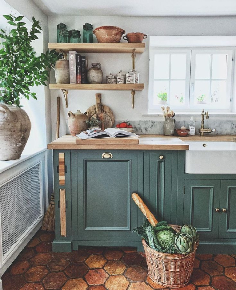 Charming rustic French farmhouse kitchen with green painted cabinets (Farrow & Ball Green Smoke), floating shelves, farm sink, and collected antique pots and baskets - Vivi et Margot. #frenchfarmhouse #rustickitchens #farmhousekitchen #greenkitchens #frenchcountry #kitcheninfrance #oldworldstyle