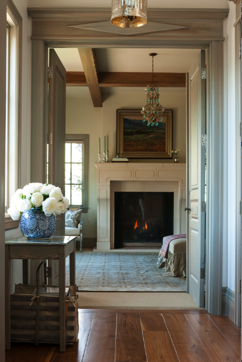 A elegant hallway with wide plank hardwood floors looking into living room with French limestone fireplace. Trim is stained in a Nordic style blue gray. Interior design by Decor de Provence.