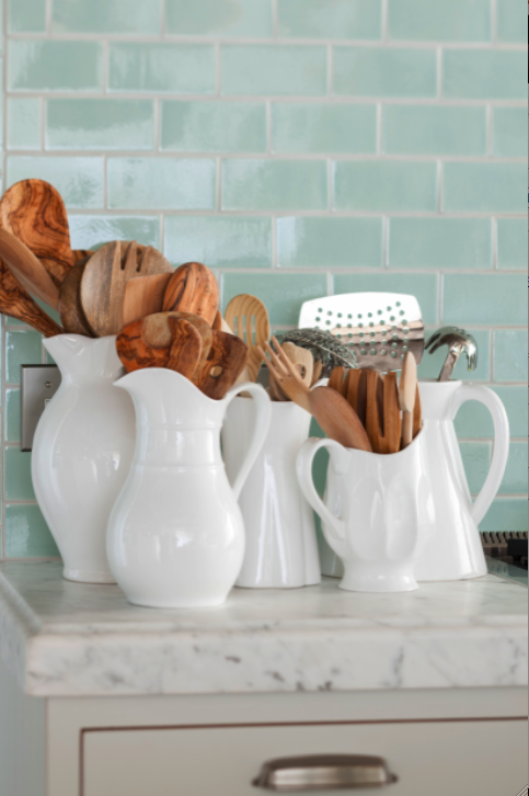 A collection of white pitchers hold wooden spoons and kitchen essentials in a French country cottage kitchen with glazed green tile. Decor de Provence. Come be inspired by interior design photos with French Green Paint Colors and Serene French Blue-Greens. #greenpaintcolors #mintgreen #interiordesign #paint