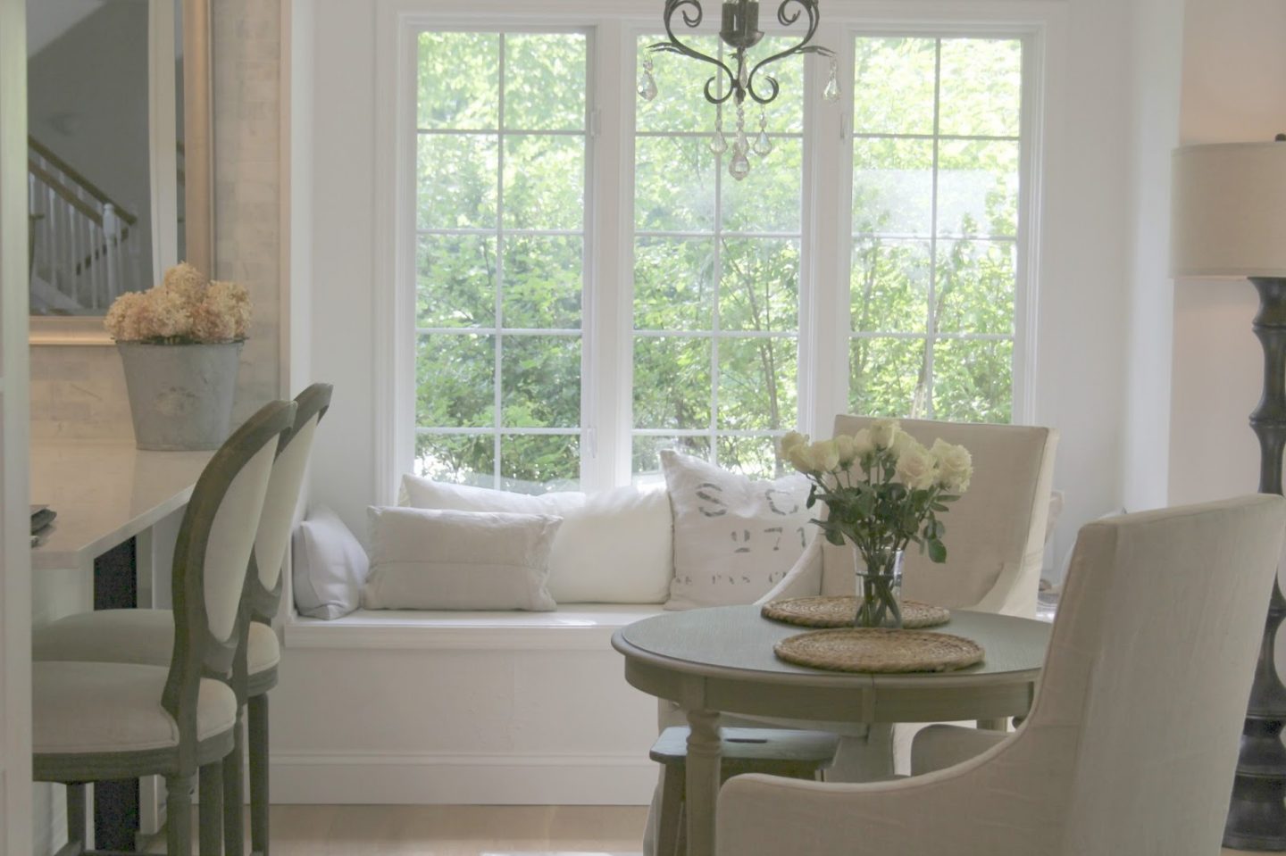 Serene whit kitchen with windowseat. Countertop is Viatera quartz in Minuet. Paint color is Benjamin Moore WHITE. Belgian linen French bar stools and slipcovered arm chairs with round wood dining table - Hello Lovely Studio. #hellolovelystudio #kitchendecor #frenchcountry #serene #belgianlinen #europeancountry #neutral #windowseat #breakfastroom