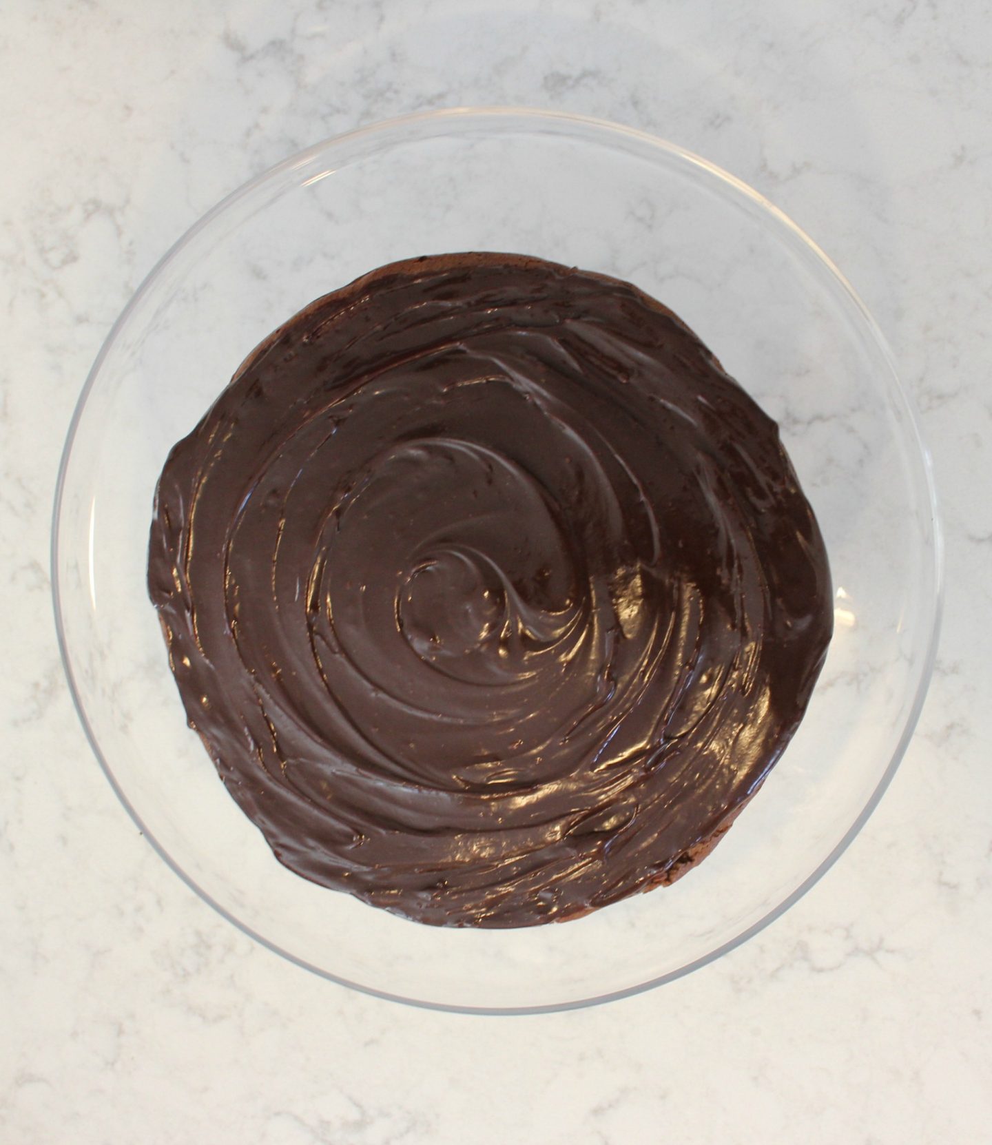 Round chocolate ganache iced cake on clear glass cake pedestal - Hello Lovely Studio