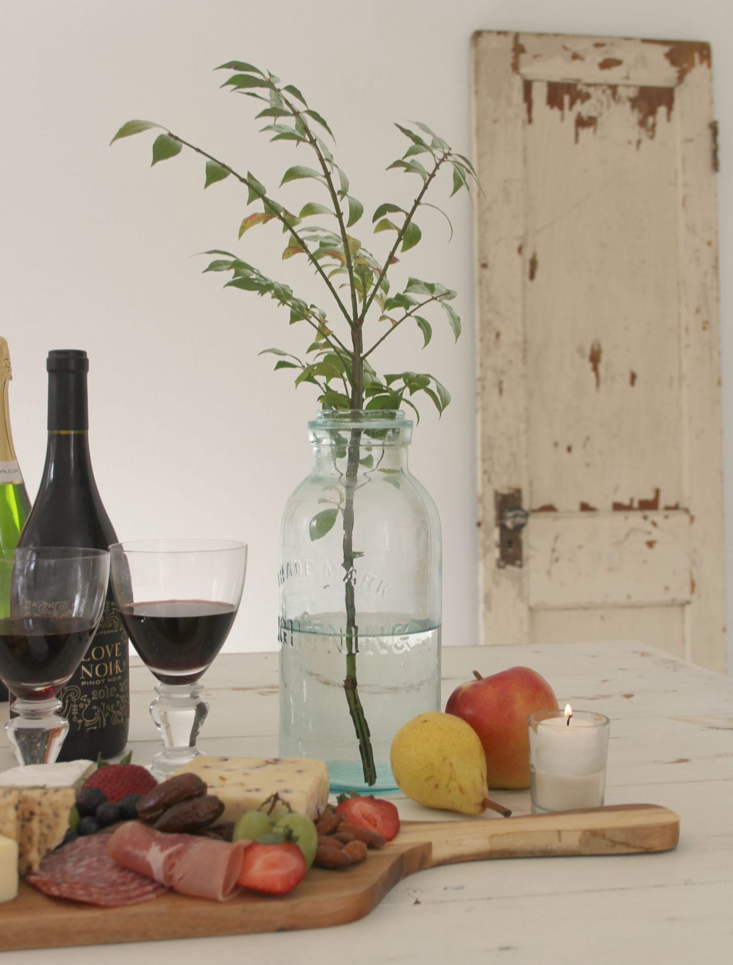 Weathered white farm table with cheeseboard, red wine, rustic branch in blue bottle, and peely shabby chic door in background. Come discover how to decorate chic yet cheap! #hellolovelystudio #shabbychic #tablescape #cheeseboard #vintagedoor