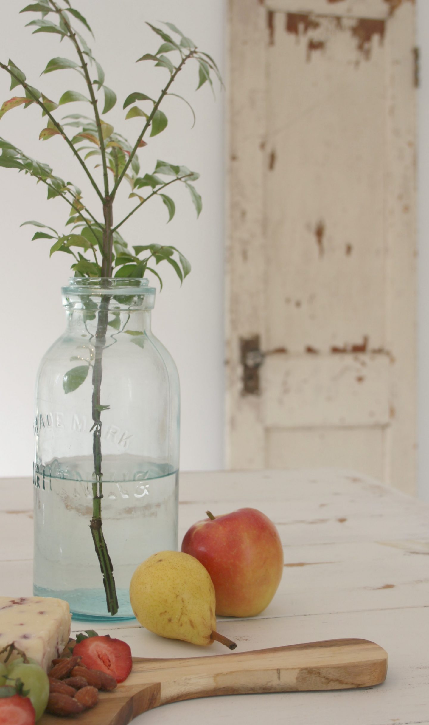 Serene vignette in my studio with white reclaimed barnwood farm table, old mason jar as vase, colorful fruit, rustic cutting board, and peely weathered vintage door in background. Come tour my home and consider inexpensive approaches to simple decor. 