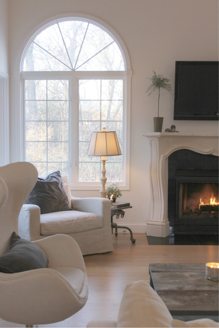 Belgian linen upholstered chairs, white oak hardwood flooring, and a French fireplace in our European inspired country living room - Hello Lovely Studio.  You'll love these white paint color ideas!  #interiordesign #paintcolors