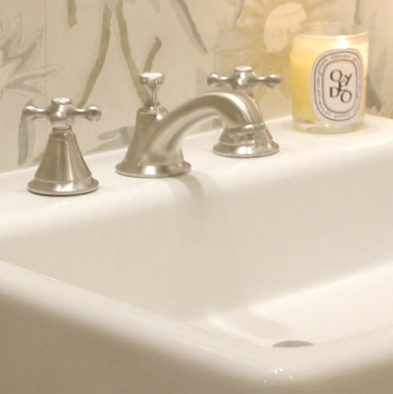 Detail of a gorgeous brushed nickel widespread Seabury bathroom faucet by Grohe. The cross handles lend a nostalgic and classic feel. A Dyptique candle glows on the farm sink. Design by Hello Lovely Studio.