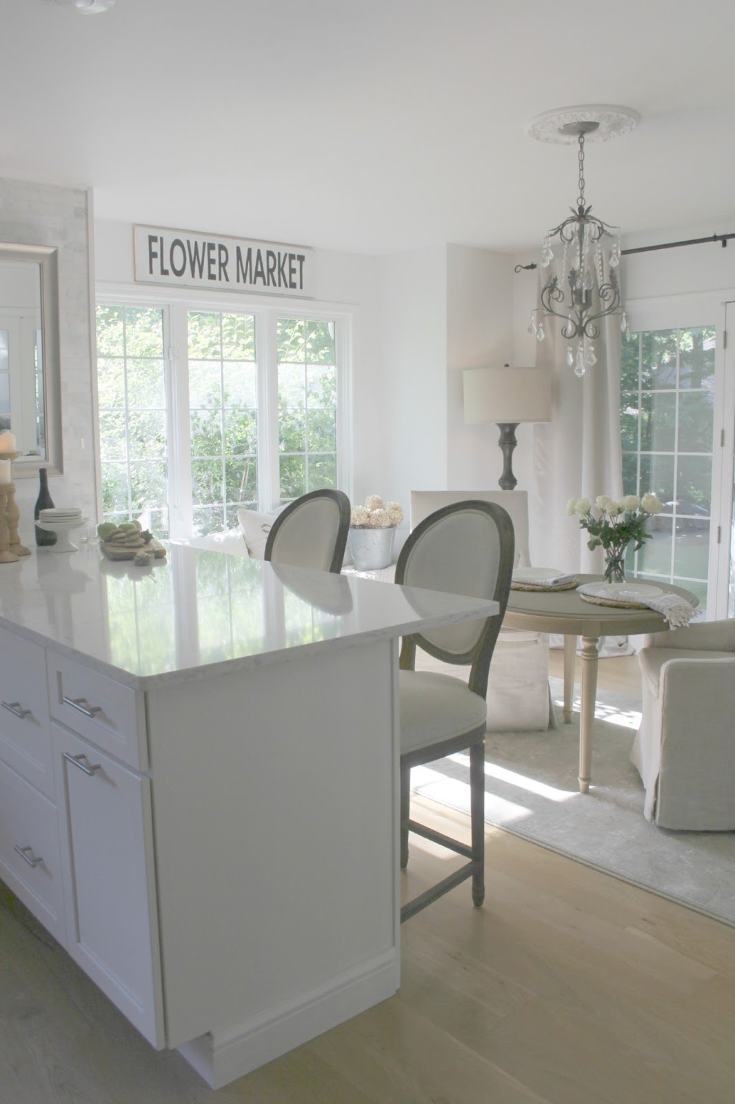 White kitchen with serene decor, Minuet quartz counters, linen chairs, vintage sign, and crystal chandelier. Design by Hello Lovely Studio. #whitekitchen #modernfarmhouse #frenchcountry #europeancountry #serene #timeless #hellolovelystudio