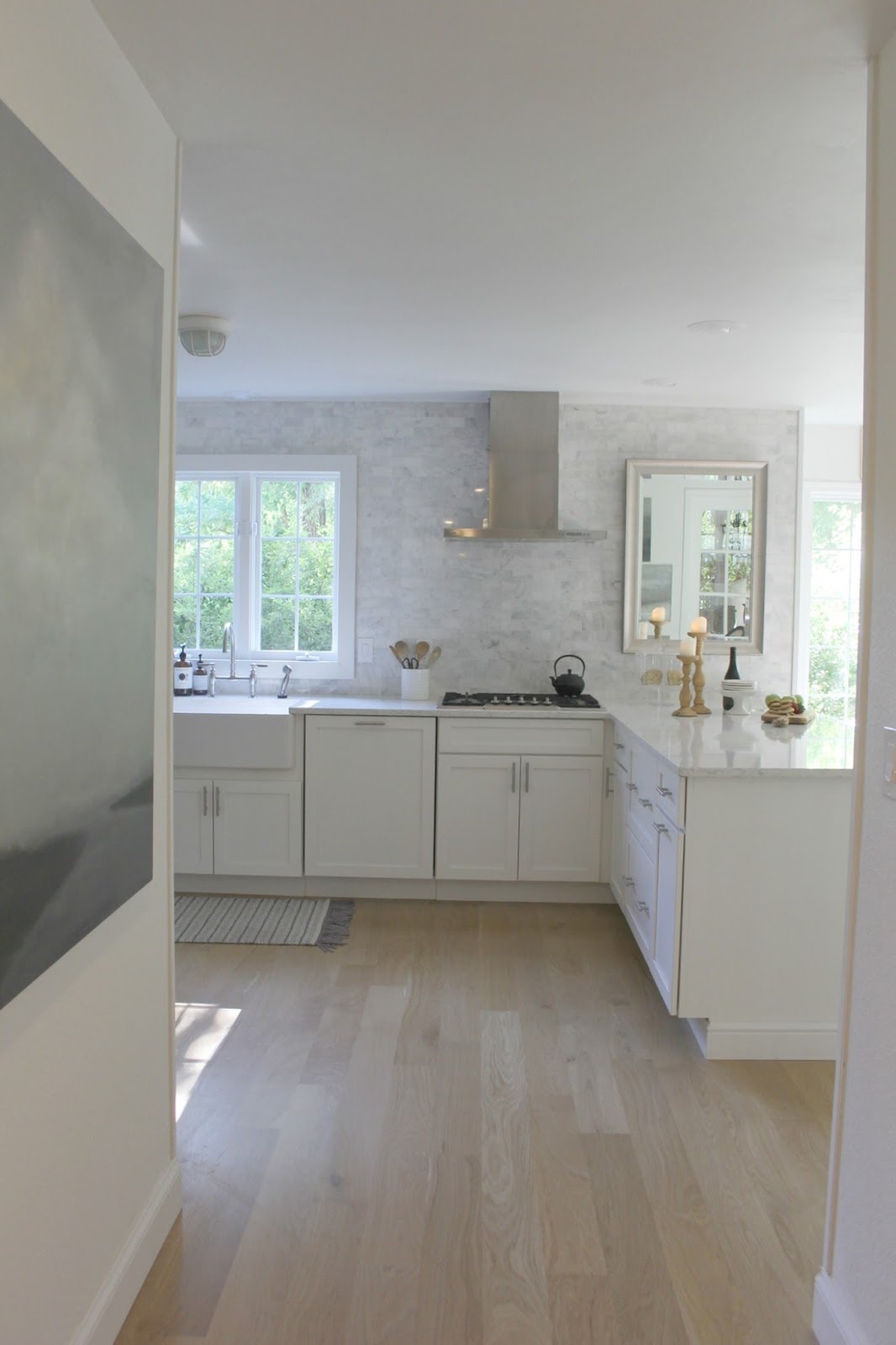 Serene white modern farmhouse kitchen with European inspired details. Shaker cabinets, marble subway tile, white oak hardwood, and abstract painting on wall. #whitekitchen #kitchendecor #modernfarmhouse #kitchenideas #serene #hellolovelystudio #whitedecor