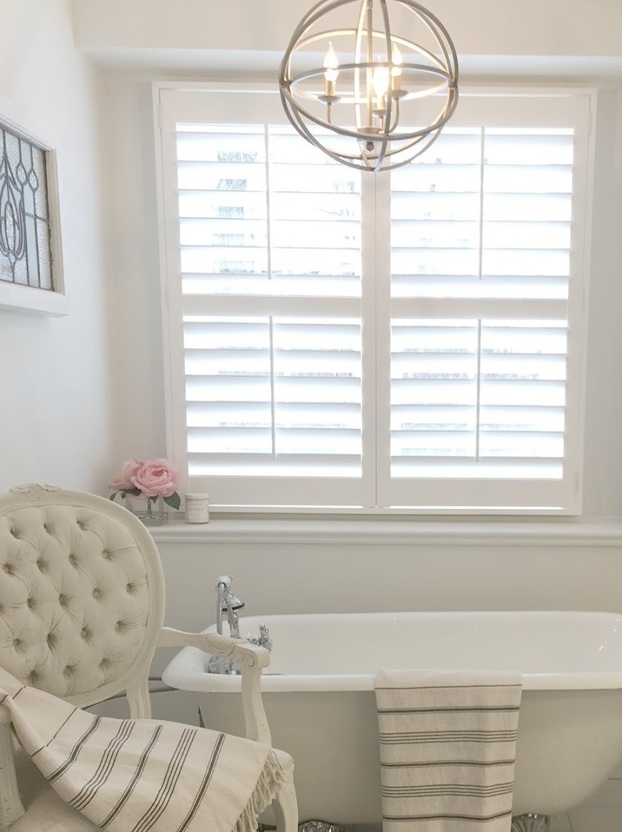 Paint color is Benjamin Moore White. French country style all white bathroom with plantation shutters, vintage clawfoot tub, tufted Louis arm chair, and leaded glass window. Design and photo by Hello Lovely Studio. #whitebathroom #frenchcountry #allwhitedecor #bathroomdesign #vintagebathroom #hellolovelystudio #benjaminmoorewhite