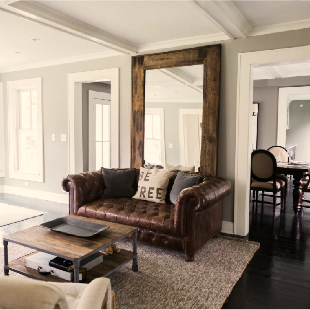 Brown leather tufted Chesterfield sofa and oversized RH rustic floor mirror in a living room with black hardwood floor.