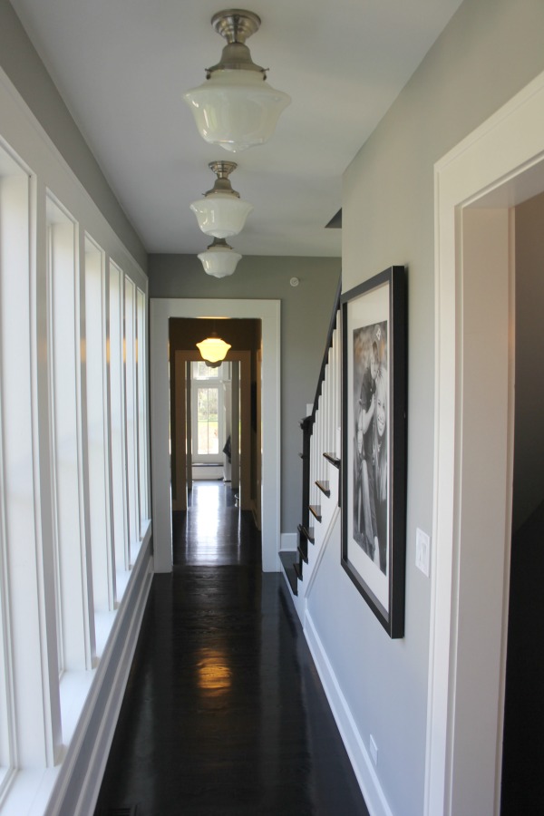 Black hardwood flooring in a hallway of a historic farmhouse with school house lights and grey painted walls. Paint color is Benjamin Moore Stonington Gray. #benjaminmoorestoningtongray #wallcolor #hallway #blackfloors #farmhouse #interiordesign #schoolhouselighting