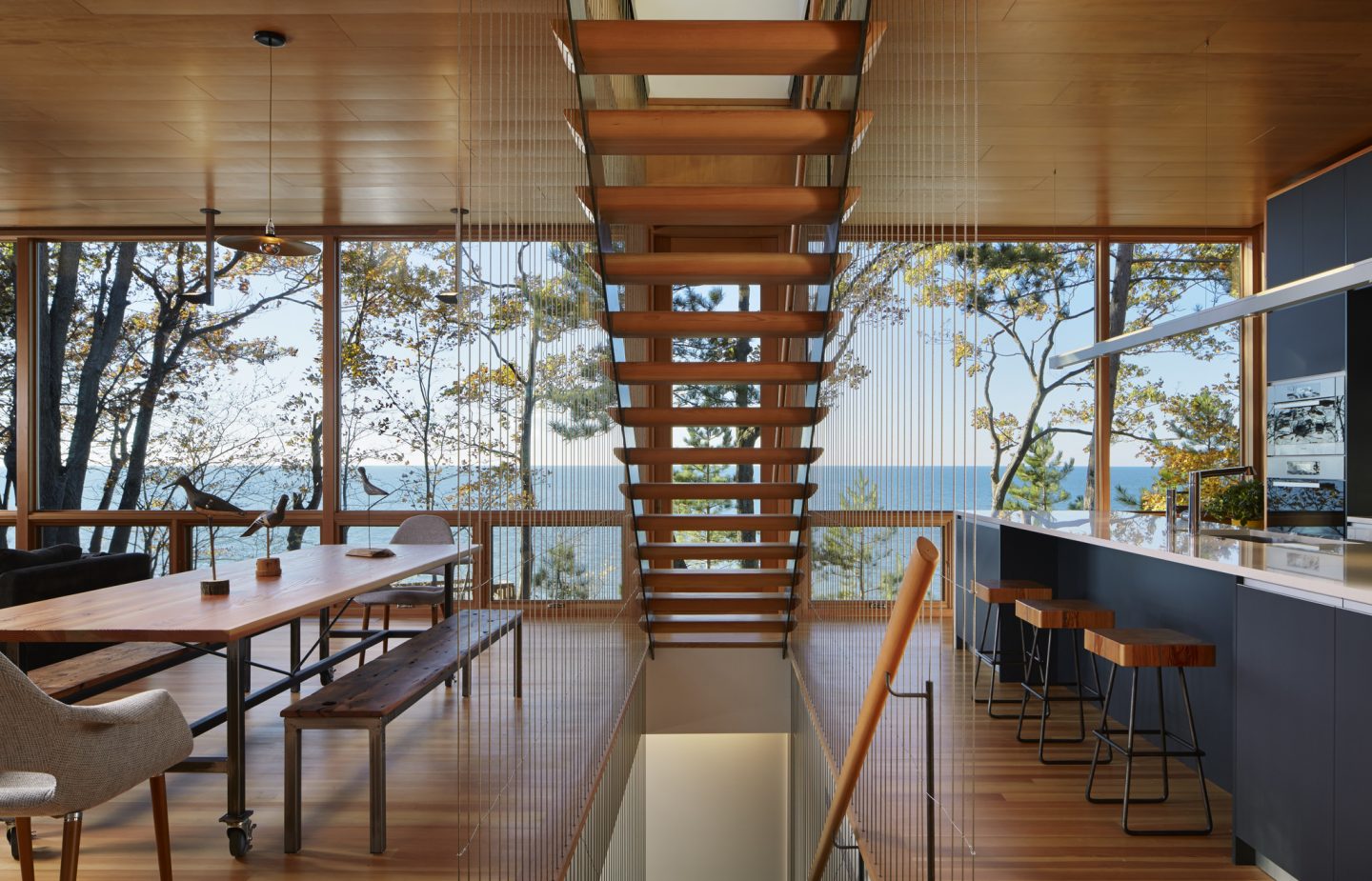 A wall of glass in a waterfront home gives an unobstructed view of nature. Modern kitchen design and open riser staircase shine in a warm yet modern house design. #waterfront #housedesign #staircase #openrisers #warmmodern