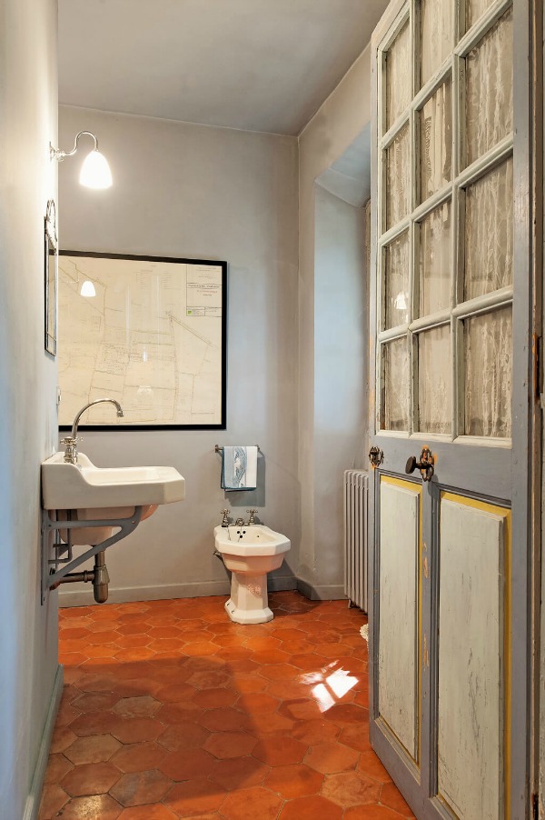 Blue grey walls and antique original door in a French country bathroom with traditional terracotta hex tile flooring and console sink - Haven In.