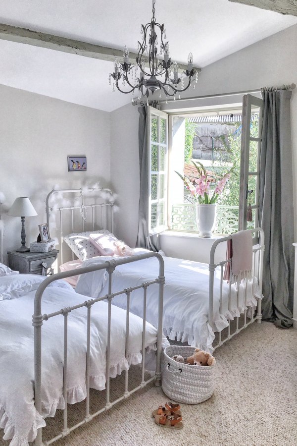 Serene, quiet, and timeless, this girls bedroom in a French farmhouse has ancient windows allowing the fresh air in, Farrow and Ball Strong White paint color on the walls, and pink accents from Pottery Barn. Vivi et Margot.