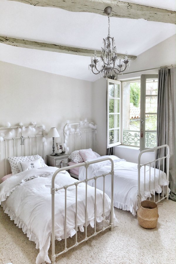 Beautiful French farmhouse bedroom with vintage style iron beds, rustic wood beams, and chandelier. Vivi et Margot. Photo by Charlotte Reiss. #vivietmargot #frenchfarmhouse #bedroom #frenchbedroom #farmhousebedroom