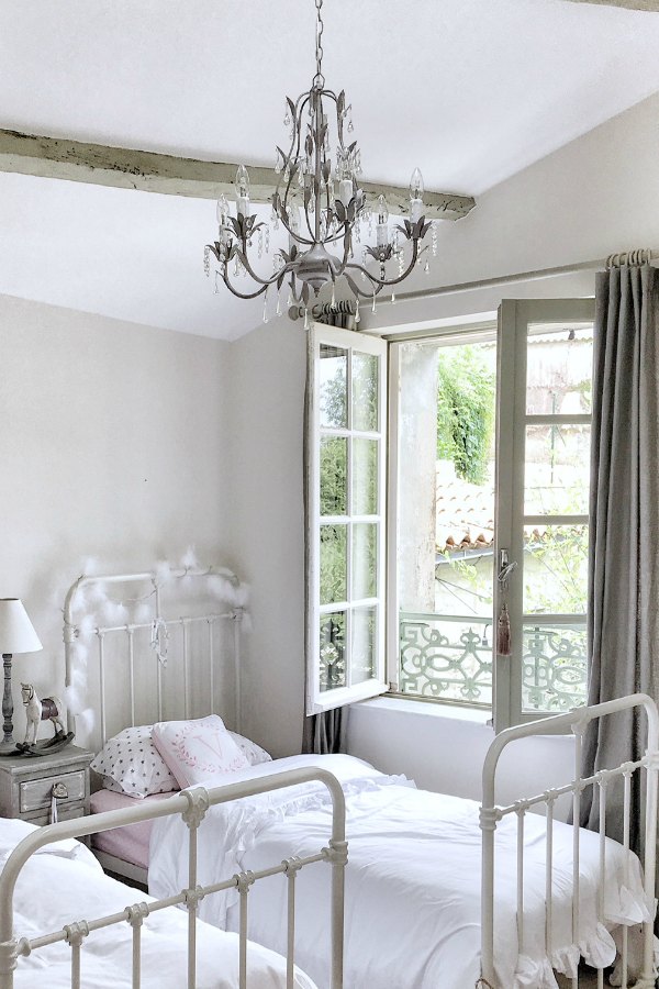 Serene, quiet, and timeless, this girls bedroom in a French farmhouse has ancient windows allowing the fresh air in, Farrow and Ball Strong White paint color on the walls, and pink accents from Pottery Barn. Vivi et Margot.