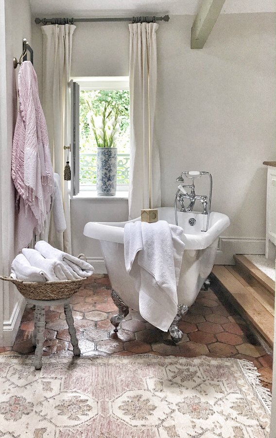 Quiet and timeless hues in a French farmhouse bathroom by Vivi et Margot features reclaimed terracotta hex tiles, freestanding tub, romantic chandelier, and Farrow and Ball's Strong White on the walls.