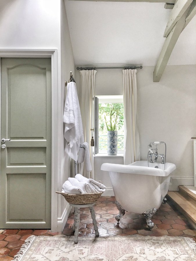 French farmhouse bathroom with Farrow and Ball French Gray painted trim, Strong White on walls, clawfoot tub, beamed ceiling, and antique terracotta floor tiles. Vivi et Margot. Find the right white paint color for your own space!