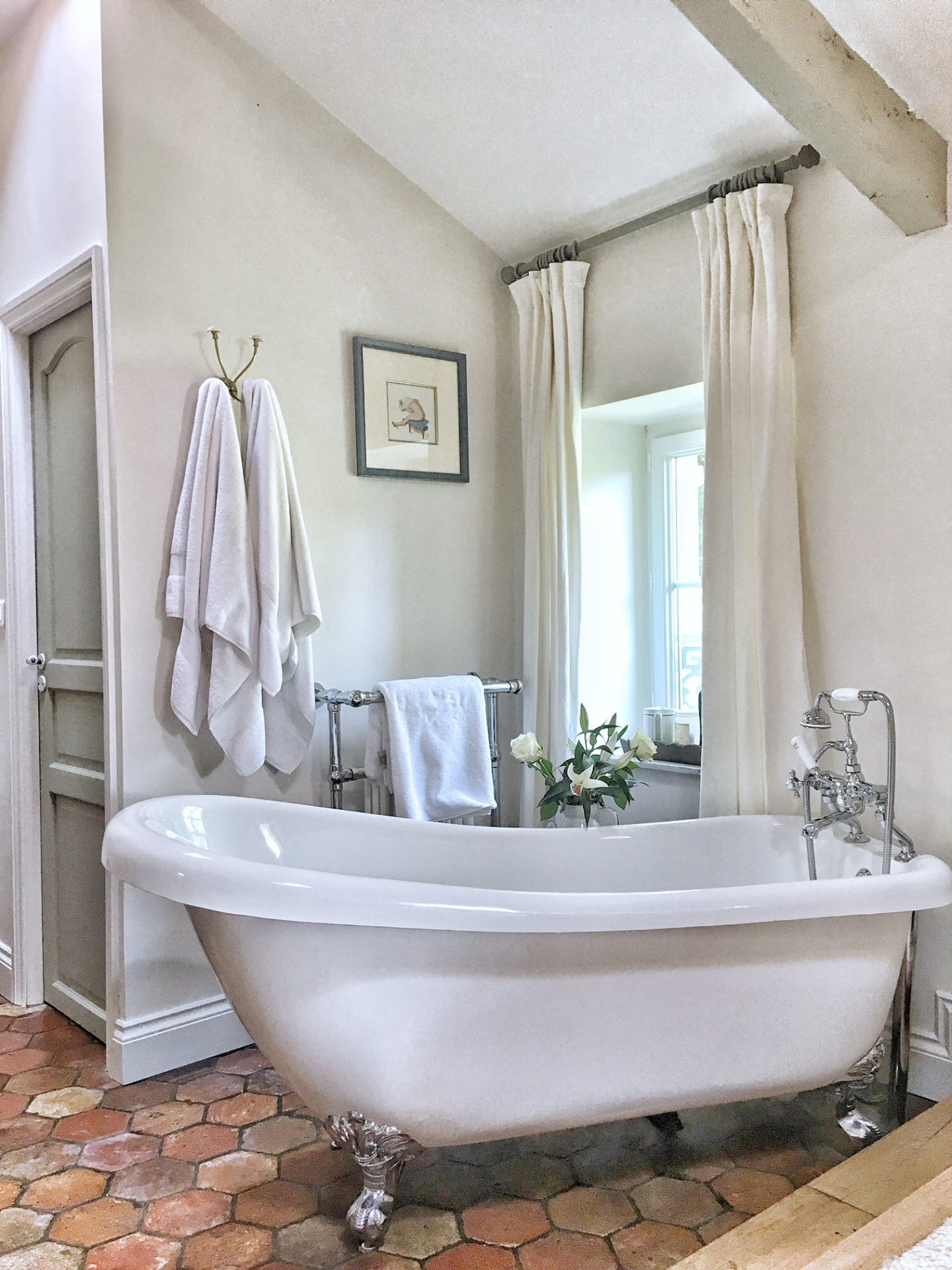 Quiet and timeless hues in a French farmhouse bathroom by Vivi et Margot features reclaimed terracotta hex tiles, freestanding tub, romantic chandelier, and Farrow and Ball's Strong White on the walls.