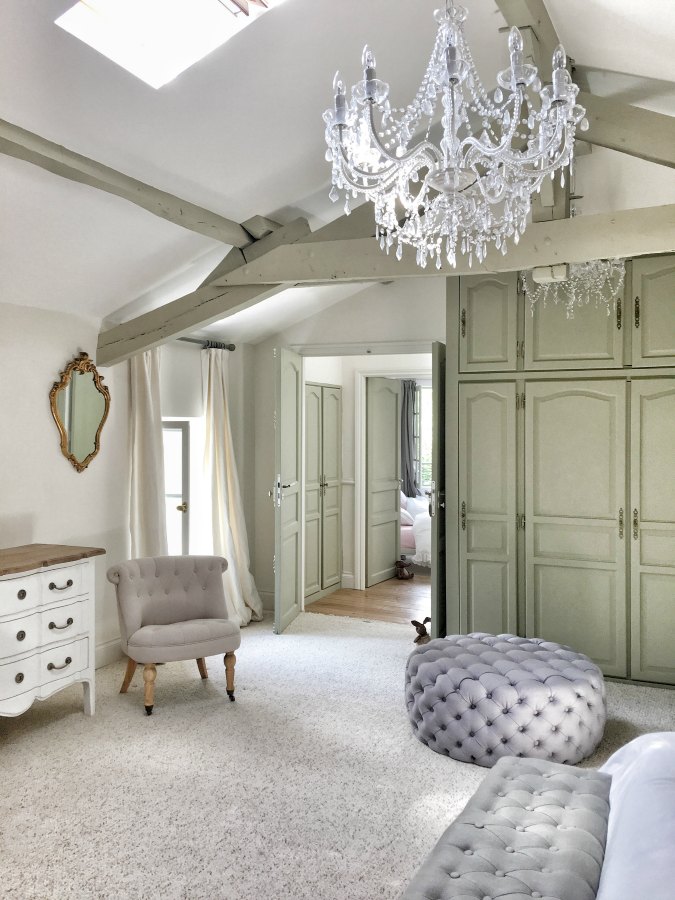 Built-ins, tufted furniture, crystal chandelier, and rustic beams in a renovated farmhouse bedroom in France by Vivi et Margot. #frenchfarmhouse #bedroomdecor #farrowandballstrongwhite #interiordesign