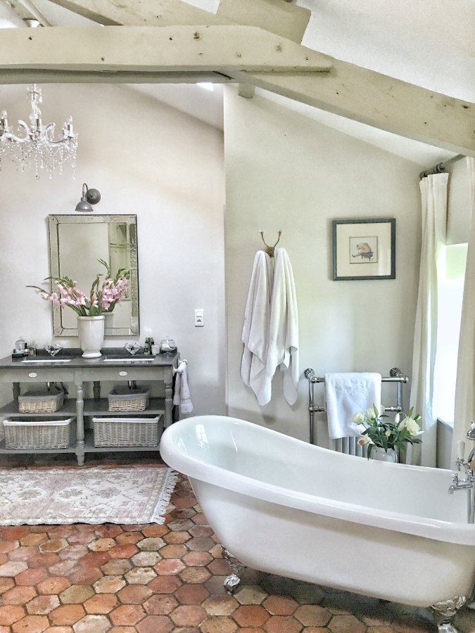 Quiet and timeless hues in a French farmhouse bathroom by Vivi et Margot features reclaimed terracotta hex tiles, freestanding tub, romantic chandelier, and Farrow and Ball's Strong White on the walls.