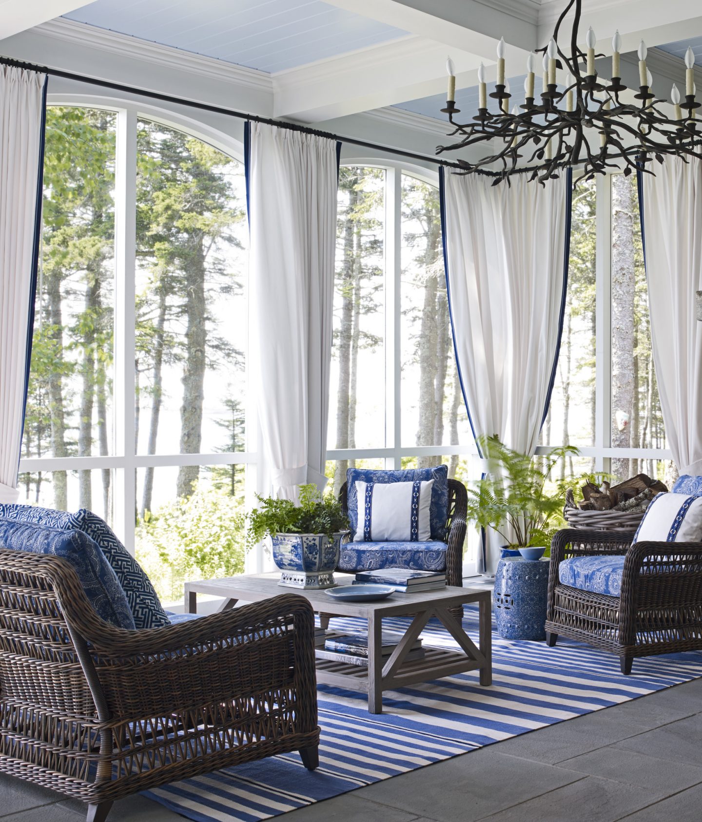 In an architecturally magnificent home in Maine with interior design by Suzanne Kasler, the enclosed porch is attached to the main house. Linen curtains give the exterior room the feeling of an interior room. Bold blue and white stripe rug, various printed fabrics on rattan chair cushions, and blue garden stools are classic coastal design elements offering a warm welcome with cool tones.