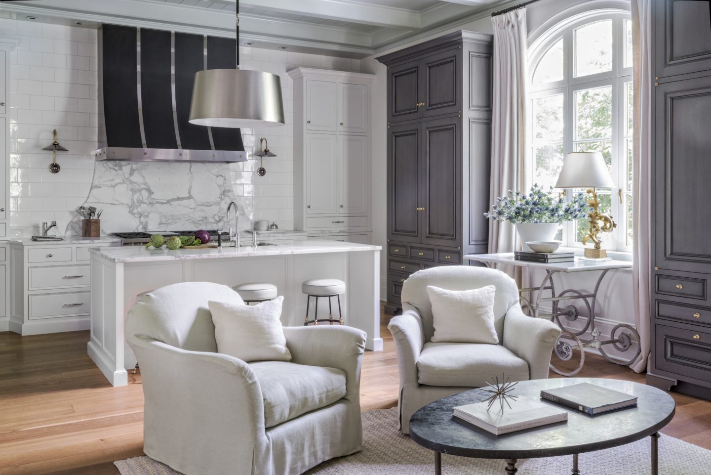 A tailored white kitchen with design by Suzanne Kasler. White walls and weathered grayarmoires. Custom black hood with metallic detail over range. The French pastry table was found at 1stdibs. Belgian chairs in rustic linen. #suzannekasler