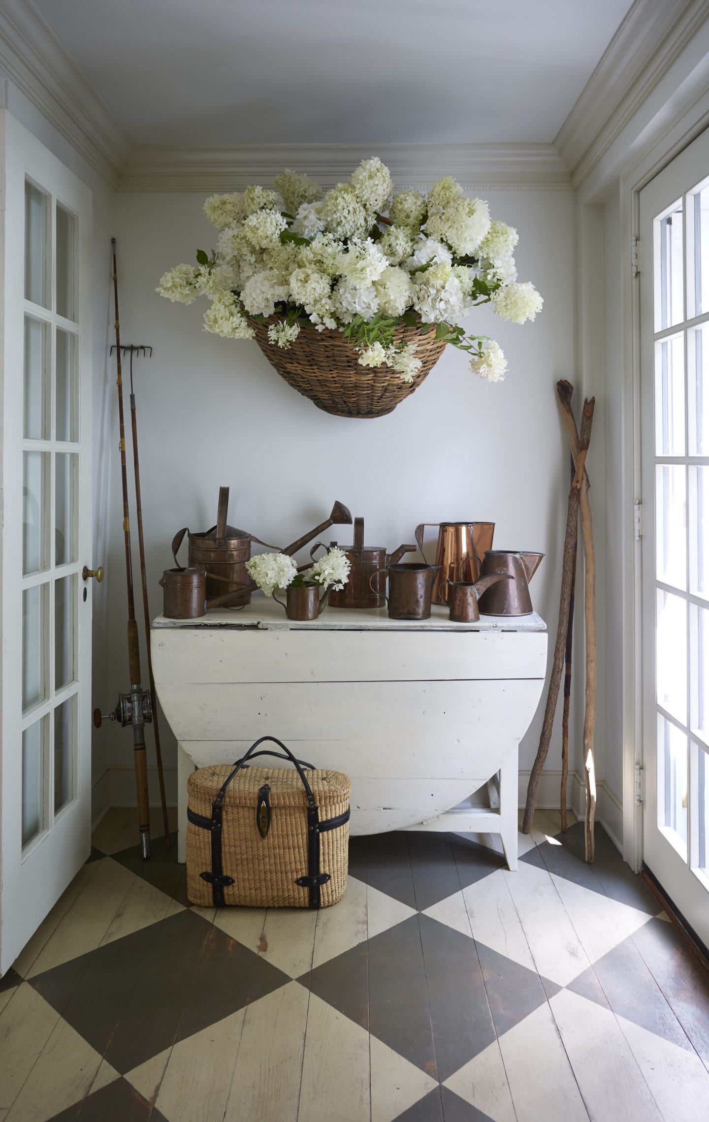 Nora Murphy country house entry with checkerboard pattern flooring, dropleaf table, and huge basket of hydrangea and collection of watering cans. Nora Murphy Country Style to Inspire! #noramuphy #countrystyle #entry #countryhouse #americanfarmhouse #farmhousestyle #rusticdecor