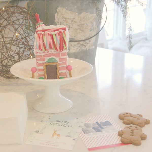 A homemade pink candy house for the holidays on a white pedestal - the siding is pink sticks of gum - Hello Lovely Studio.