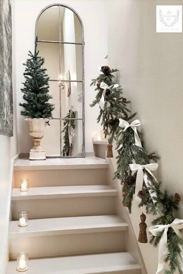 White Christmas decor in a French country home with minimal simple beauty on a staircase with fresh greenery, white bows, and pale decor. Beautiful holiday decor inspiration from interior designers and bloggers. Come discover 28 Amazing Christmas Decorating Ideas! #christmasdecor #holidaydecorating