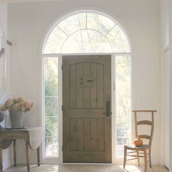 Knotty rustic alder wood planked front door with speakeasy in a lofty entry with antique dropleaf table, dried hydrangea, and French prayer chair  within a European Country cottage by Hello Lovely Studio.