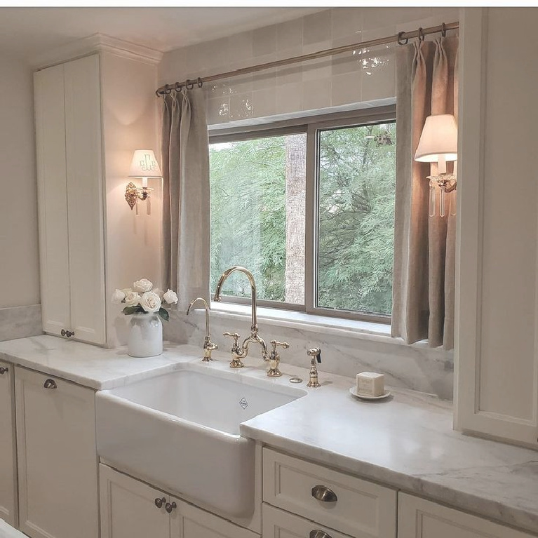 Romantic all white French country kitchen by French Nest Interior Design Co with farm sink flanked by sconces, linen curtain panels, and simple classic cabinetry. #frenchcountry #kitchens #frenchkitchens #kitchendesign #farmsink #romanticdecor