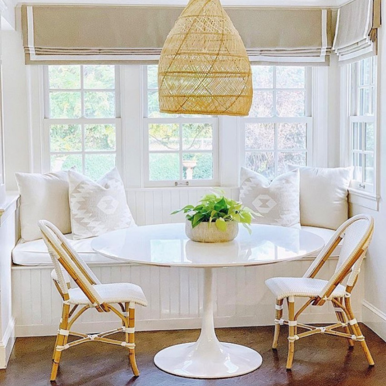 Lovely coastal white kitchen breakfast nook with Saarinen style table, banquette, wicker pendant, and Parisian bistro chairs from Serena & Lily. #breakfastnook #banquette #whitekitchen #windowseat #serenaandlily #saarinentable