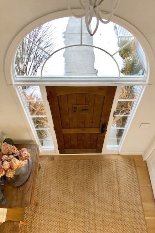 Rustic alder front door in an entry with jute area rug. Hello Lovely Studio. Walls are painted Benjamin Moore White.