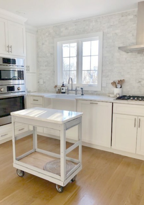 Classic white coastal style kitchen with polished marble backsplash statement wall, Shaker cabinets, industrial work table, and Viatera quartz (Minuet). Hello Lovely Studio.