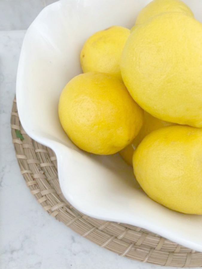 Beautiful vibrant yellow lemons in a scalloped white bowl on a round rattan charger. Hello Lovely Studio.