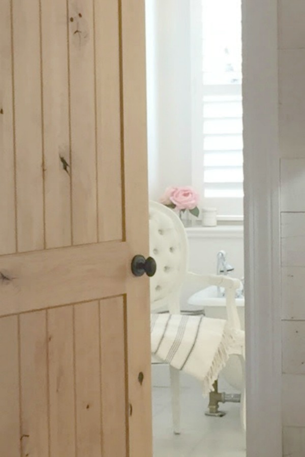 Knotty alder door leading to white French country cottage style bathroom by Hello Lovely Studio.