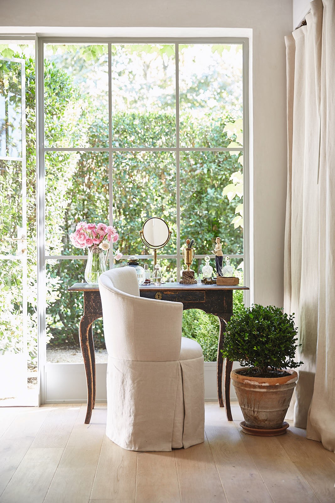 Patina Living photo of makeup area of master bathroom with antique vanity, linen upholstered chair, and linen panels. French farmhouse decor and rustic modern farmhouse style. Brooke Giannetti and Steve Giannetti designed the space at Patina Farm. Photo by Victoria Pearson.