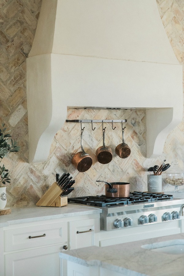 Rustic elegant French country farmhouse kitchen with beautiful stucco range hood, copper pots, reclaimed Chicago brick backsplash, arabescato marble counters, and lanterns over island. Brit Jones Design.