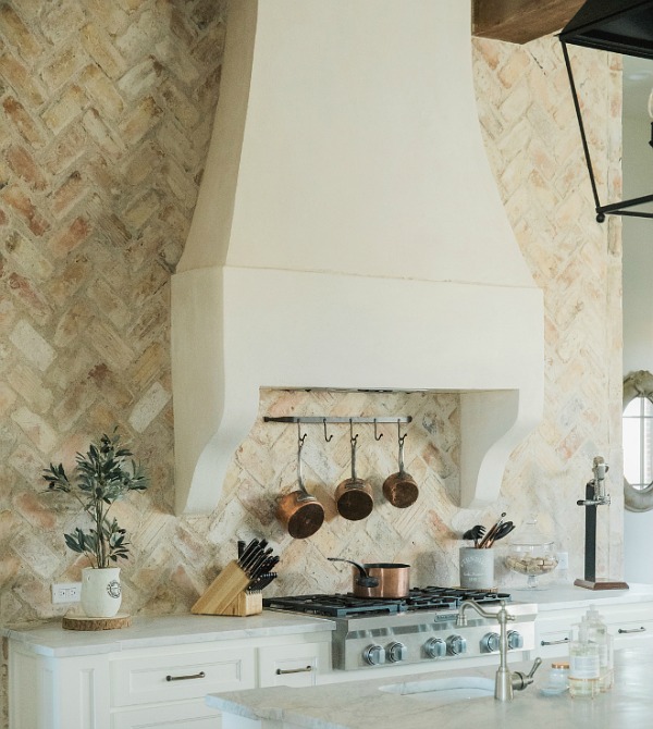 Rustic elegant French country farmhouse kitchen with beautiful stucco range hood, copper pots, reclaimed Chicago brick backsplash, arabescato marble counters, and lanterns over island. Brit Jones Design.