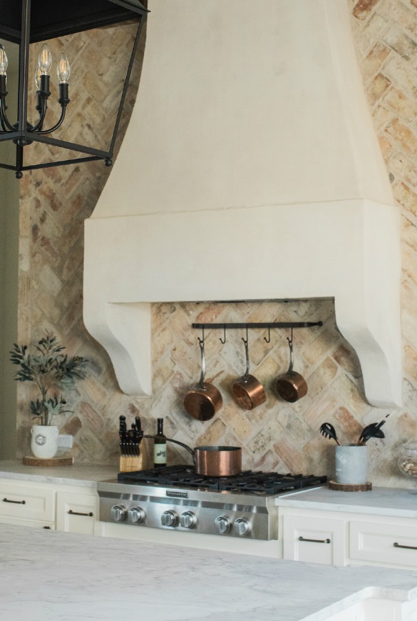 Rustic elegant French country farmhouse kitchen with beautiful stucco range hood, copper pots, reclaimed Chicago brick backsplash, arabescato marble counters, and lanterns over island. Brit Jones Design.