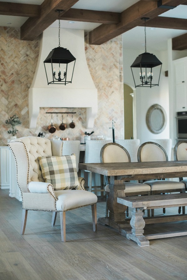 French country farmhouse kitchen with wingback chair, French dining chairs, farm table, rustic hanging lantern pendants, and reclaimed Chicago brick. Brit Jones Design. Come see 24 Inspiring European Country Kitchen Ideas!