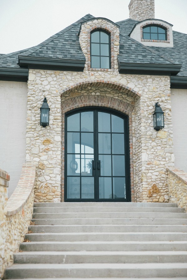 Elegant French country home exterior with steel double arch front doors, stone, brick, stucco, and rustic lanterns. Design by Brit Jones.