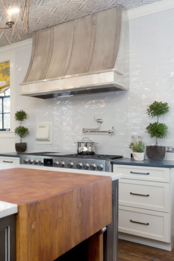 Beautiful design details in the renovated white classic Julian Price House kitchen with subway tile, farm sink, and yellow accents. Marsh Kitchen & Bath executed the lovely design which features black windows, warm wood floors, butcher block insert in huge island, and luxury range and hood.