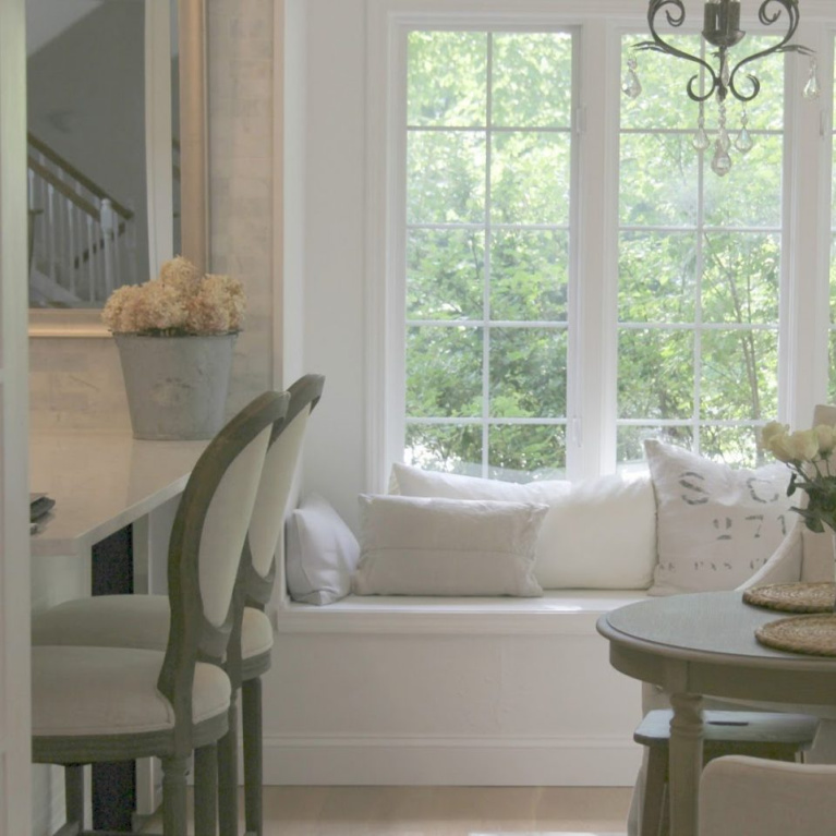 Window seat with European inspired pillows and Belgian linen slipcovered dining chairs in a breakfast area of a French country kitchen - Hello Lovely Studio. Find a Soft, Ethereal European Country Kitchen Mood to Inspire Now!