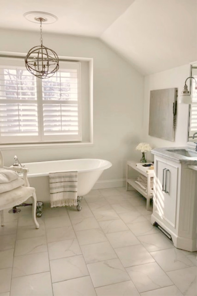 Serene white French country bathroom with vintage clawfoot tub and calacatta look porcelain tile. #hellolovelystudio #whitebathrooms #bathroomdesign #frenchcountry #clawfoottub