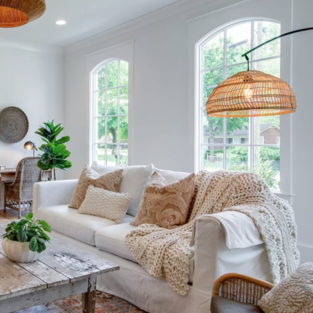 Comfy white sofa with gold pillows, cableknit chunky throw, and rattan lighting accents in a rustic cottage den in Tennessee.