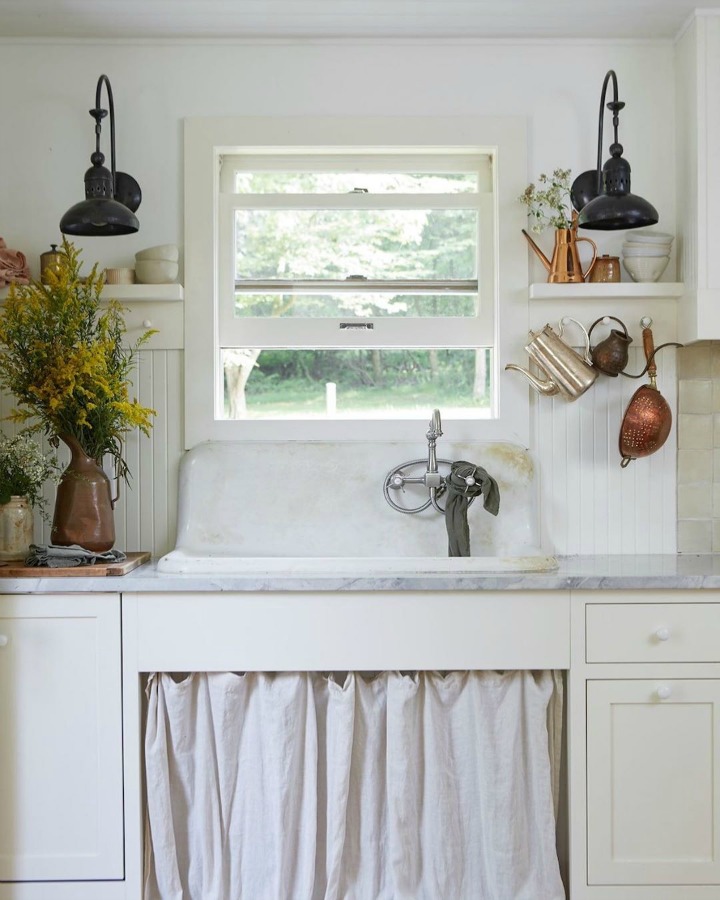 Rustic and vintage style are mellowed with white paint, a soft skirt at the sink, and edgy barn style sconces flanking the window. Design by Leanne Ford and photo by Reid Rolls for HGTV Restored by the Fords. #rustickitchen #vintagestyle #leanneford #restoredbythefords