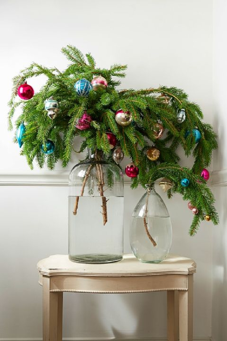 Charming and whimsical farmhouse Christmas decor with droopy evergreen branches decorated with colorful bulbs in simple white bottle vases.