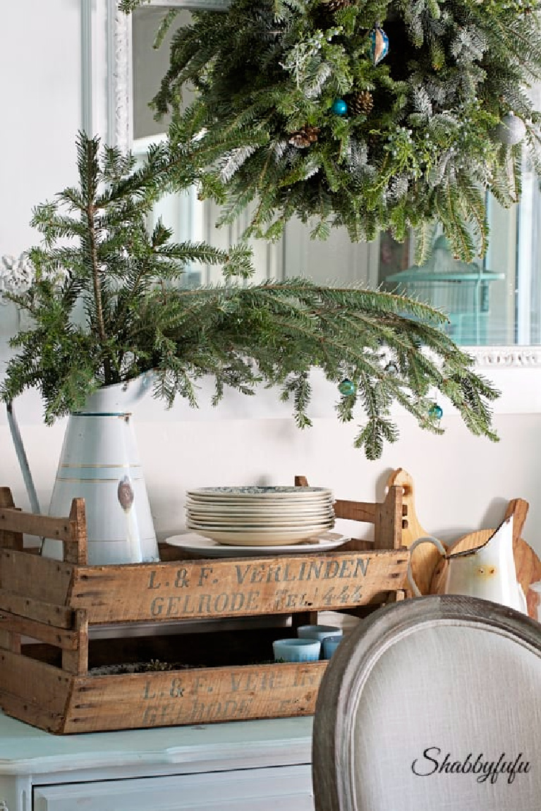 Fresh evergreen branches in a vintage enamel pitcher and rustic wood box make a charming vignette for a Christmas dining room by Shabbyfufu.