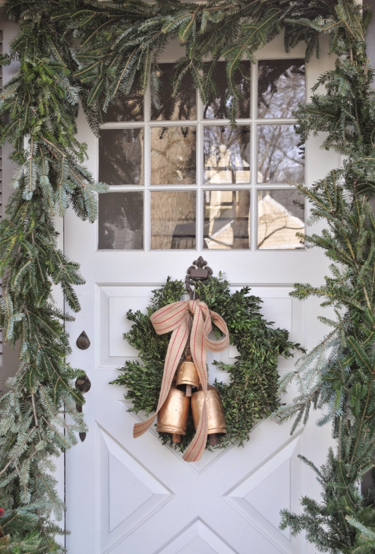 Charming and rustic Christmas wreath with gold toned bells and ribbon upon a white country door framed with fresh greenery - NineandSixteen.